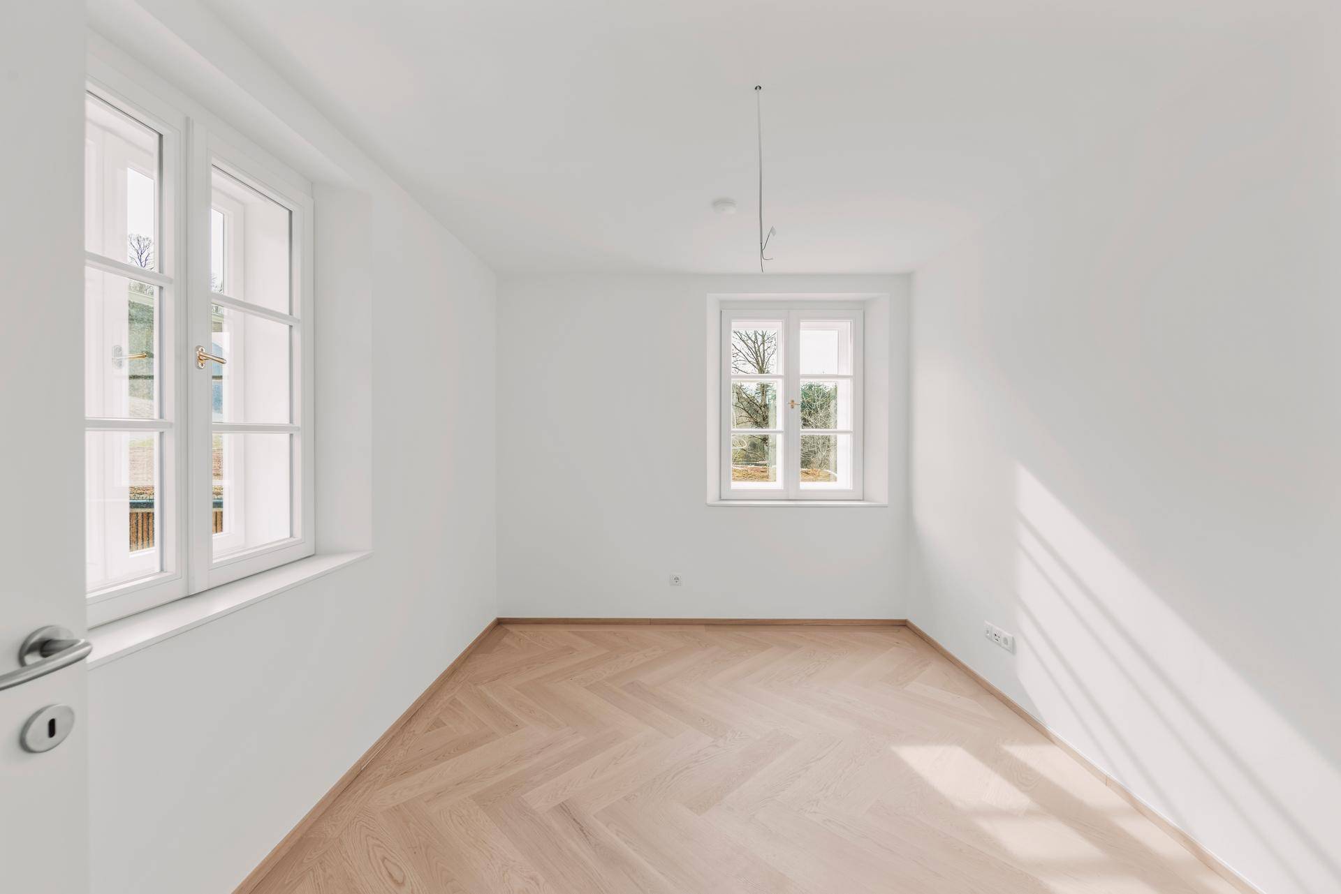 Leeres Schlafzimmer mit Fischgrätparkett, zwei Fenstern und viel Tageslicht.