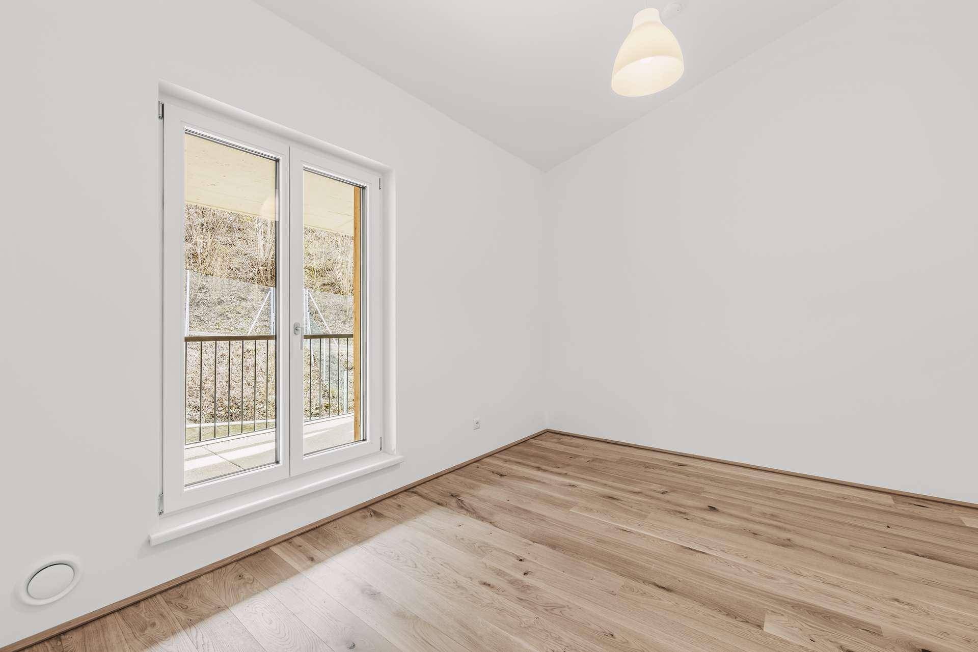 Helles, leeres Schlafzimmer mit Holzboden und Fenster mit Blick ins Grüne.