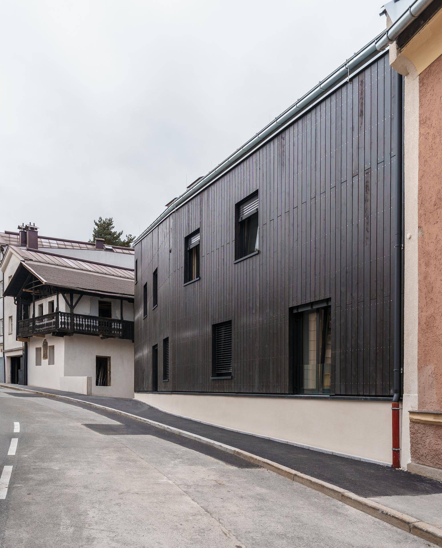 Straßenansicht des modernen Gebäudes mit dunkler Holzfassade, eingebettet in eine traditionelle Umgebung.