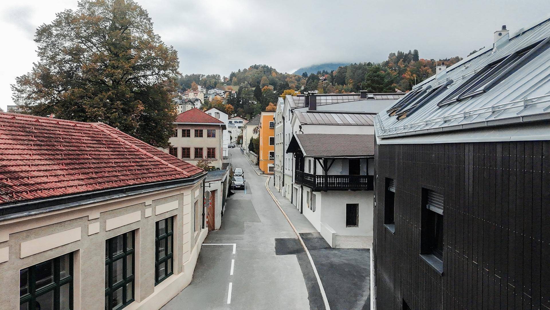 Straßenansicht von oben mit Blick auf eine Gasse, verschiedene Gebäude und herbstliche Hügel im Hintergrund.