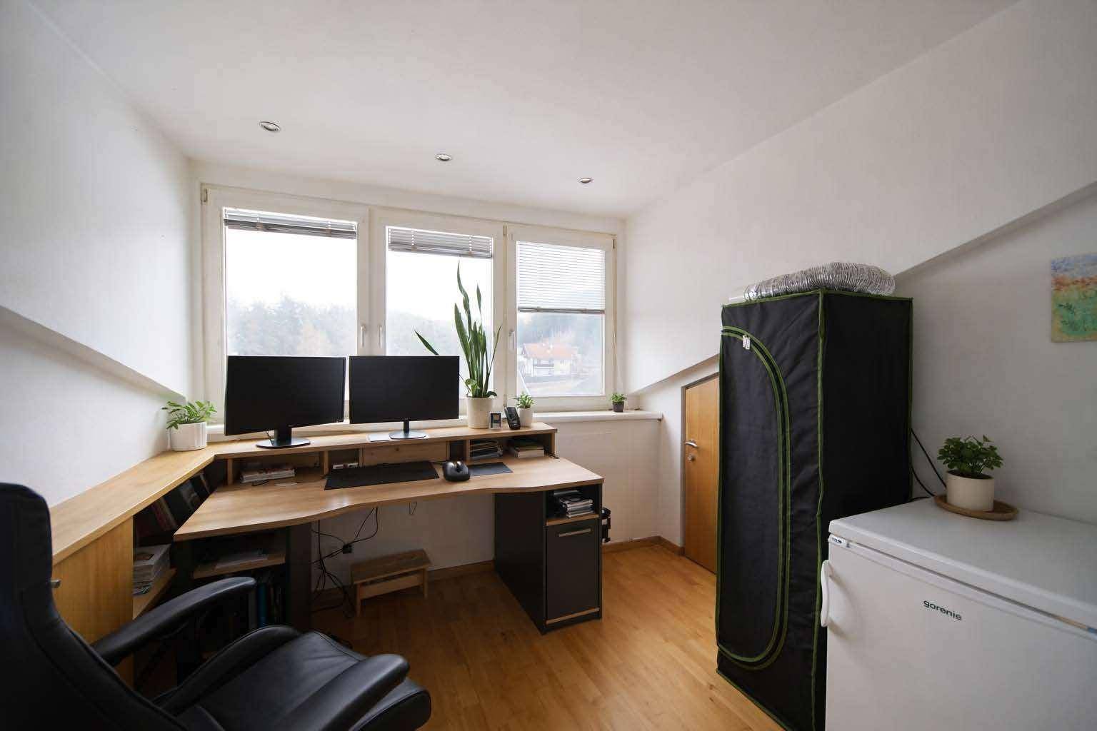 Helles Arbeitszimmer mit großem Schreibtisch, zwei Monitoren und Fensterblick unter der Dachschräge.
