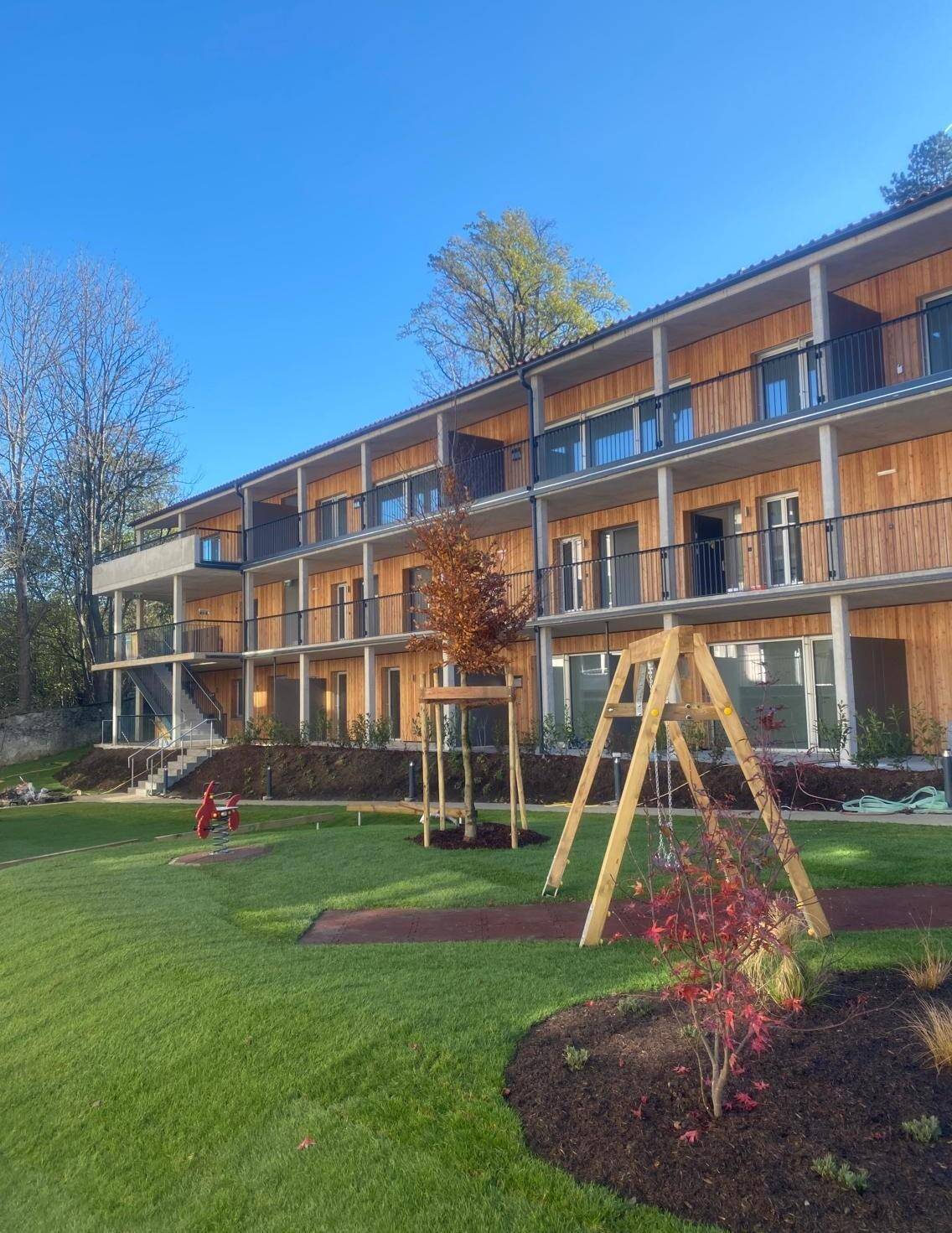 Modernes Mehrfamilienhaus mit Holzfassade, Balkonen und einem Spielplatz im gepflegten Gartenbereich.