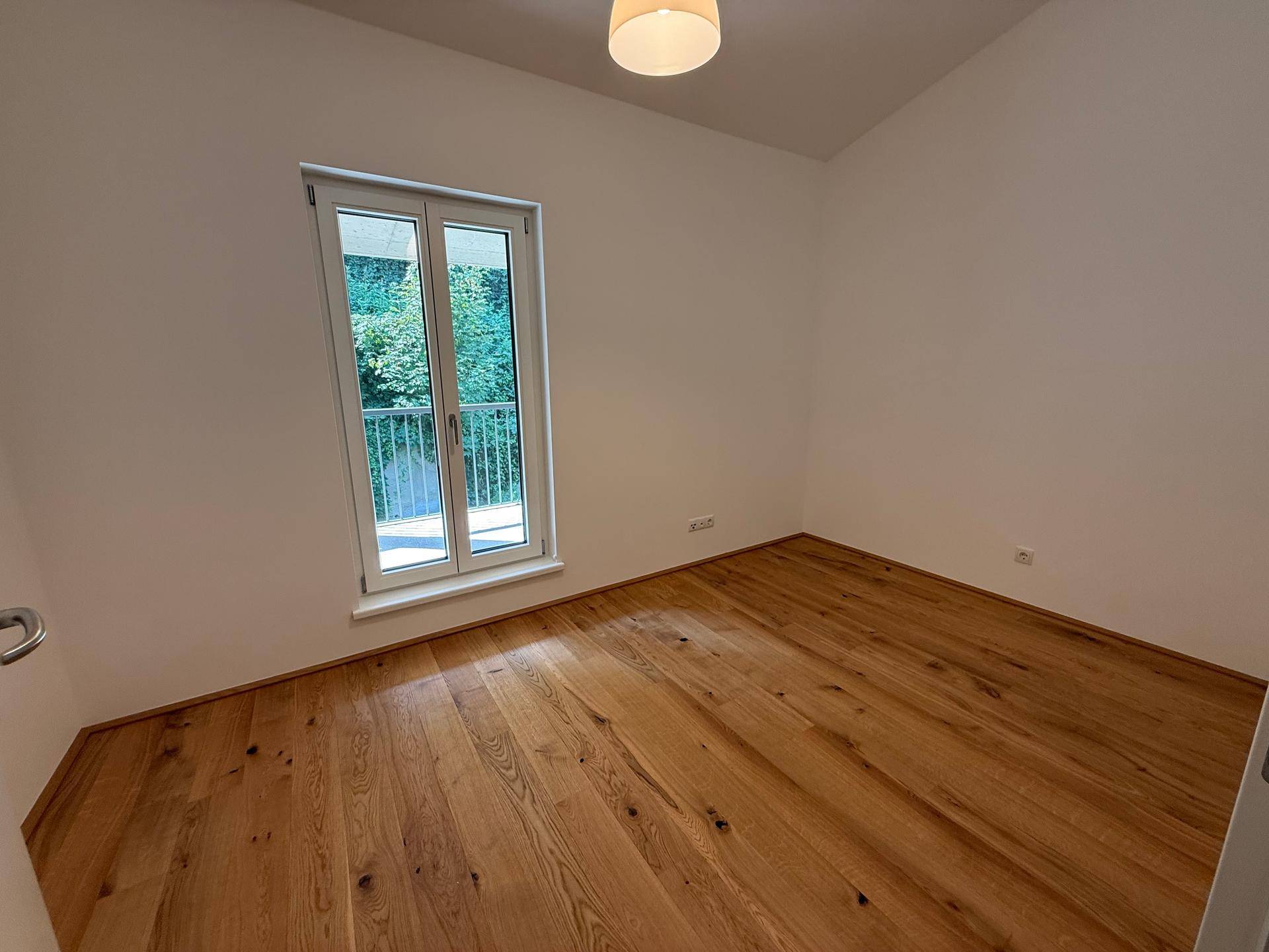 Leeres Zimmer mit Holzboden und großem Fenster, das viel Tageslicht hereinlässt.