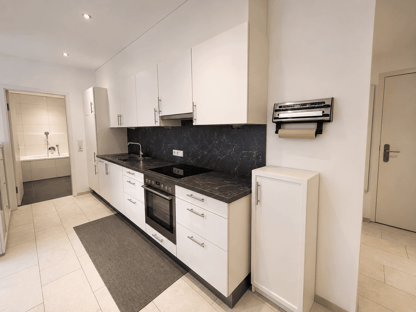 Funktionale Küche mit weißen Fronten, dunkler Arbeitsplatte und Zugang zum Badezimmer.