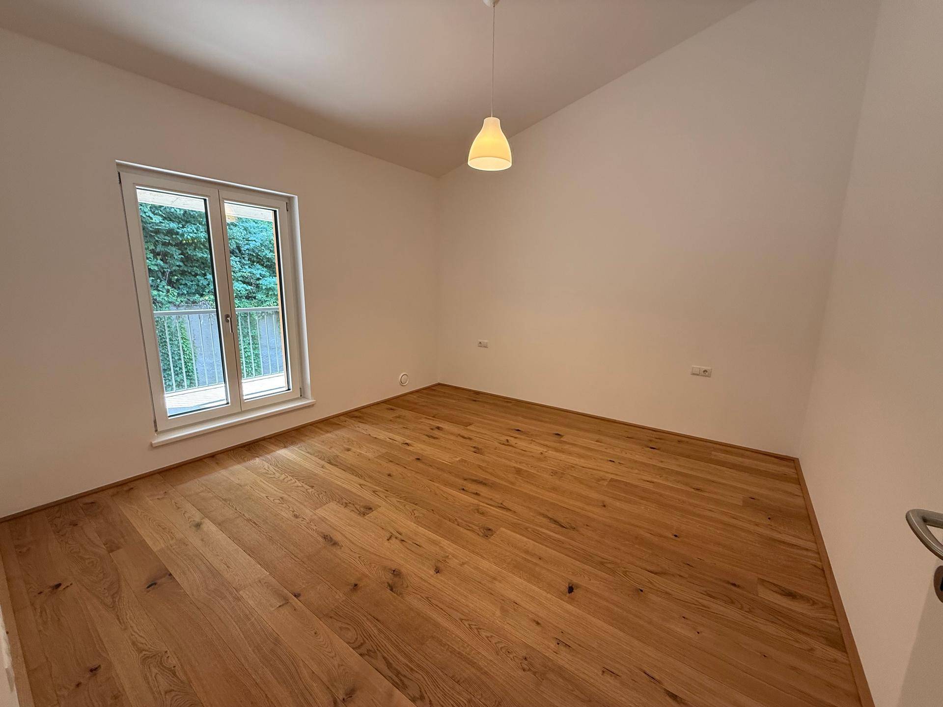 Schlafzimmer mit Holzboden, weiß gestrichenen Wänden und einem Fenster mit Blick ins Grüne.