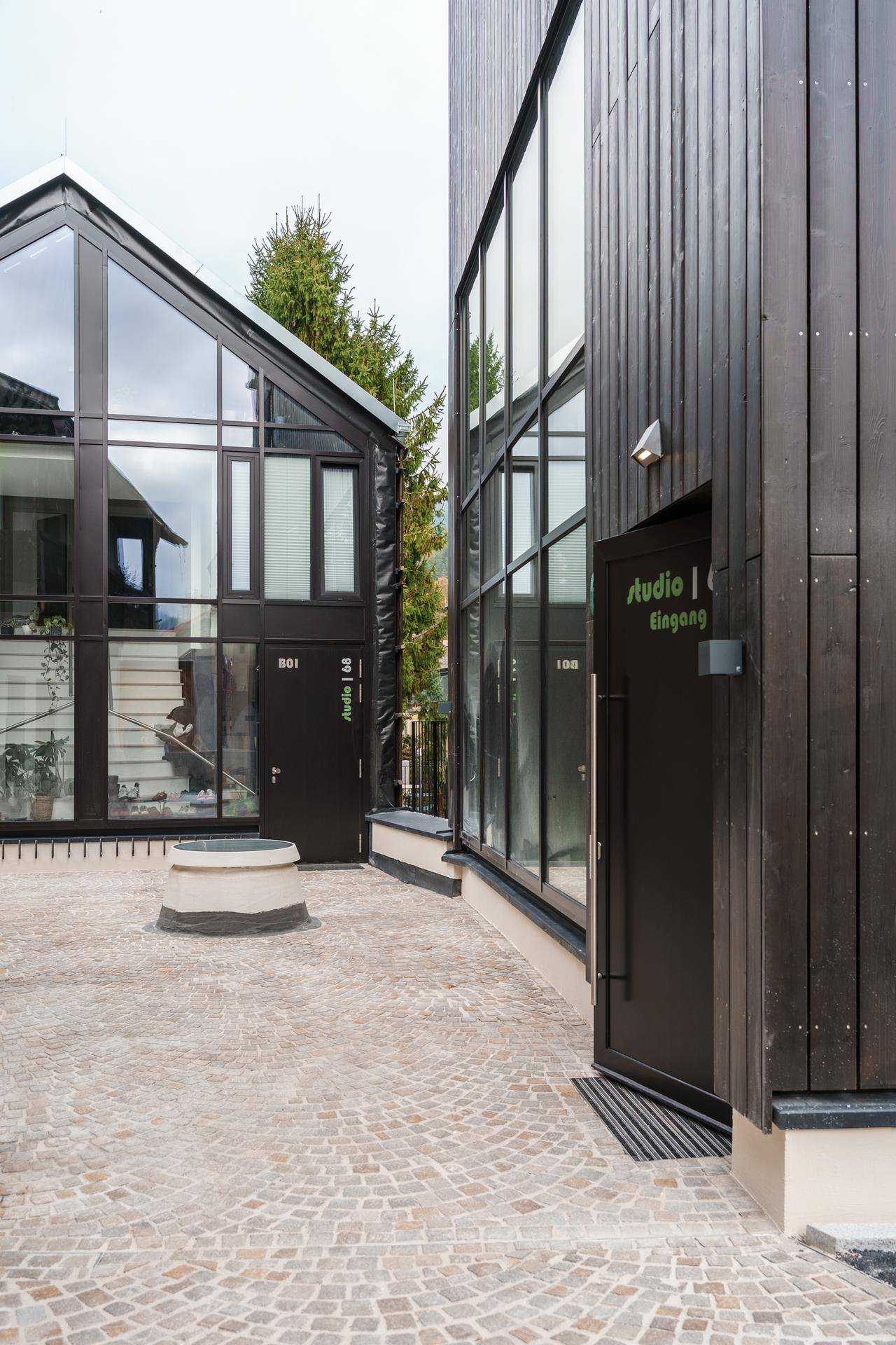 Eingangsbereich zwischen zwei modernen Gebäuden mit dunkler Holzverkleidung und großen Glasflächen.