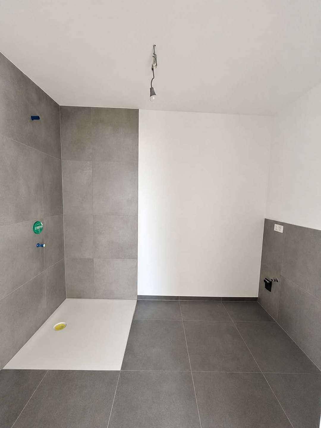 Modernes Badezimmer im Rohbau mit grauen Fliesen und vorbereiteter Duschfläche.
