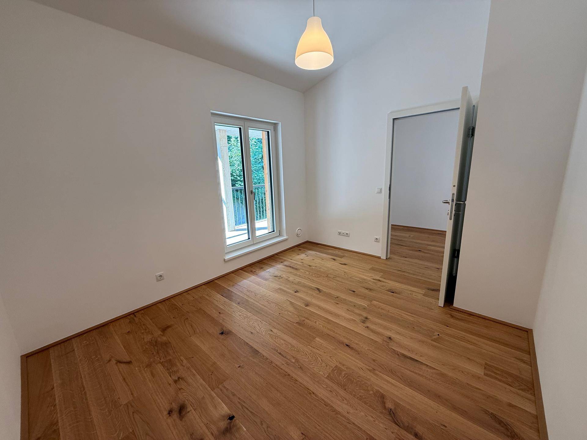 Helles Zimmer mit Holzboden und Fenster, das einen Blick ins Grüne bietet.