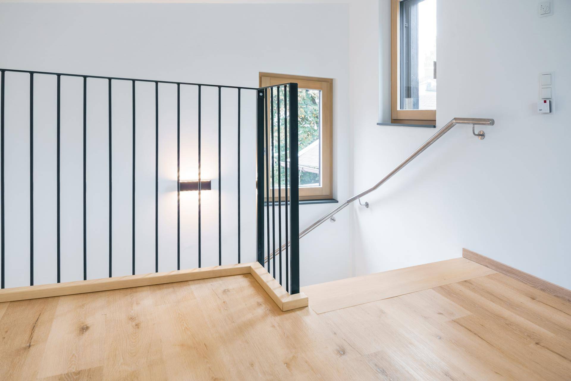 Heller Treppenabsatz mit Holzfußboden, schwarzem Metallgeländer und Fenster.