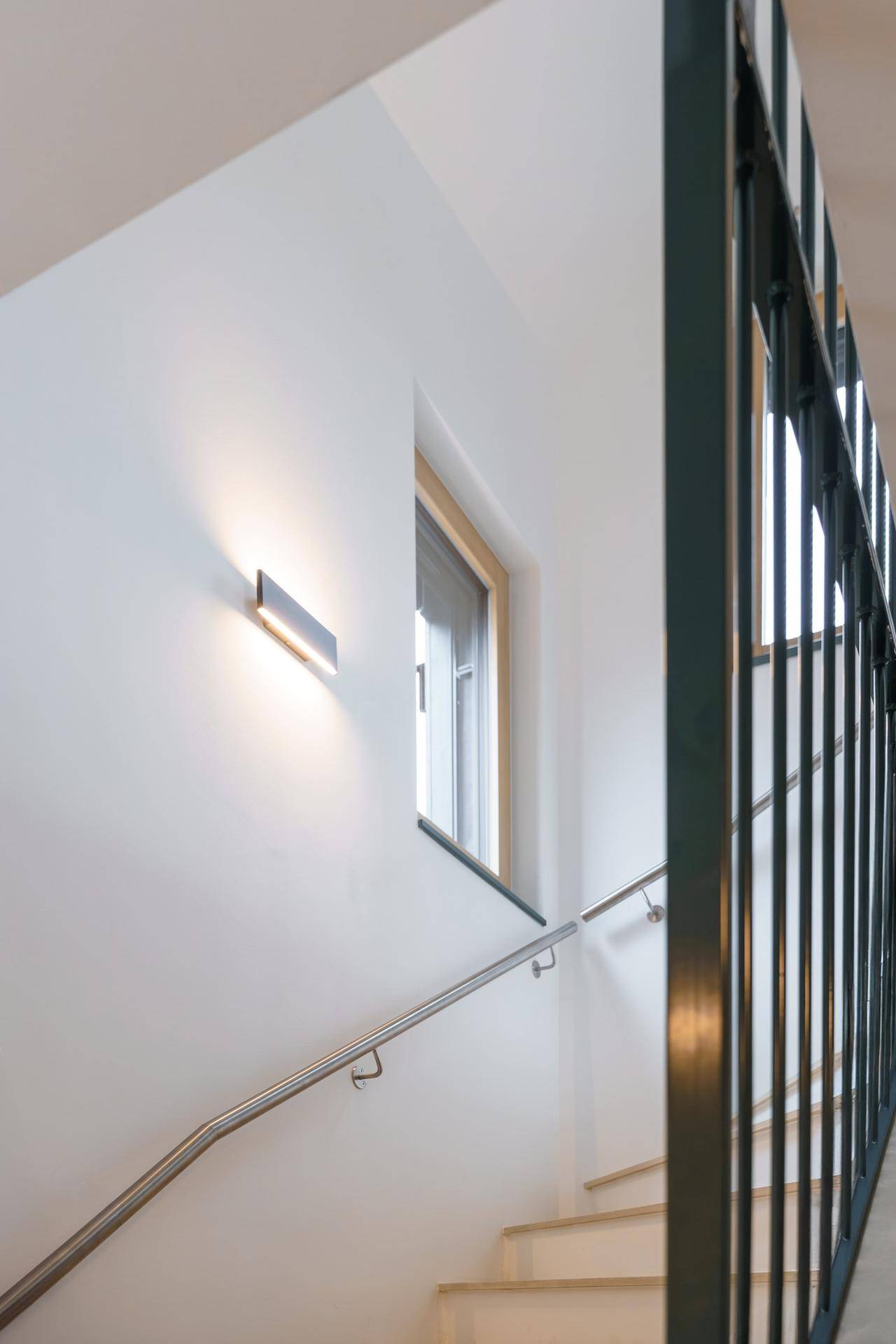 Treppenhaus mit Fenster, Edelstahlhandlauf und schwarzem Metallgeländer, beleuchtet von einer Wandleuchte.