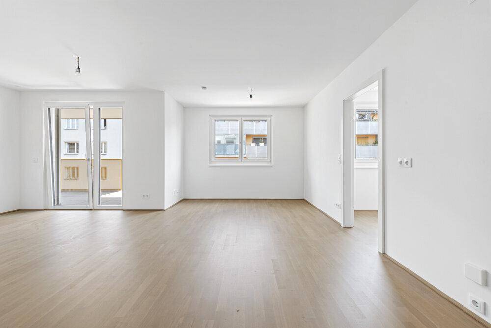 Heller, leerer Wohnraum mit hellem Holzfußboden, großen Fenstern und Balkonzugang.