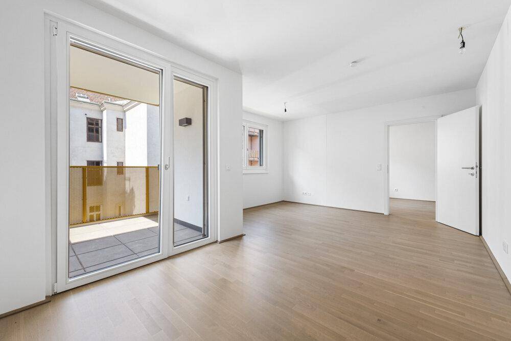 Heller, leerer Wohnraum mit Holzboden und großem Fenster mit Zugang zum Balkon.