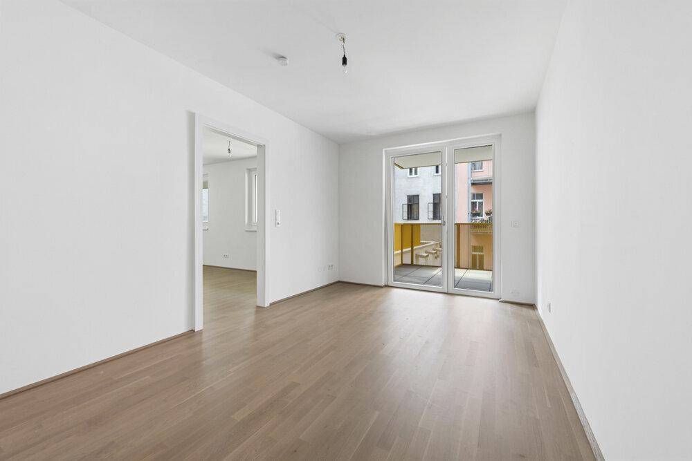 Leerer Wohnbereich mit Parkettboden und Zugang zu einem Balkon, der viel Licht hereinlässt.