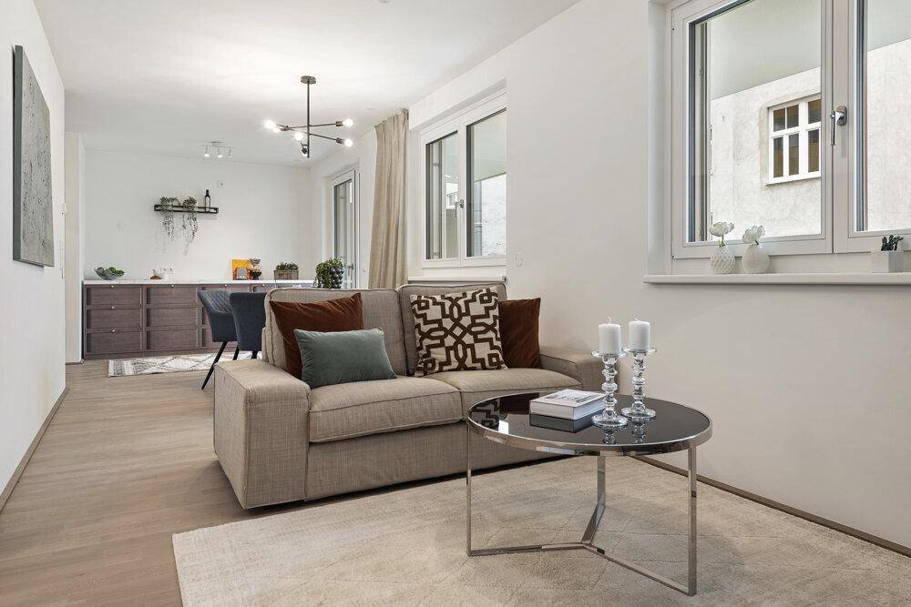 Moderner Wohnbereich mit beigem Sofa, Couchtisch und Essbereich im Hintergrund, hell durch große Fenster.