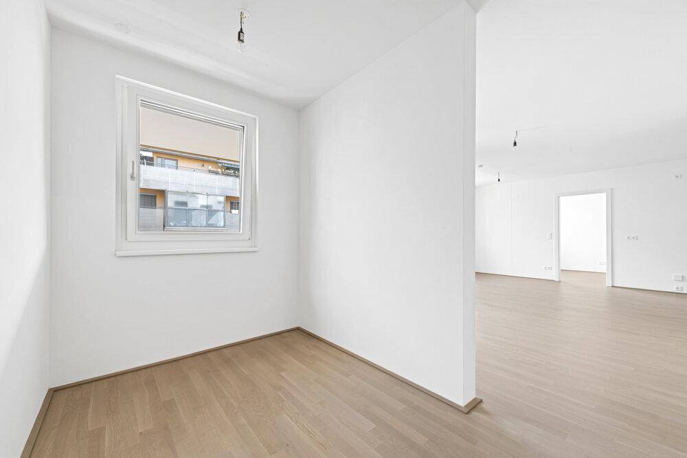 Helles, leeres Zimmer mit Parkettboden und einem Fenster, das Tageslicht hereinlässt.