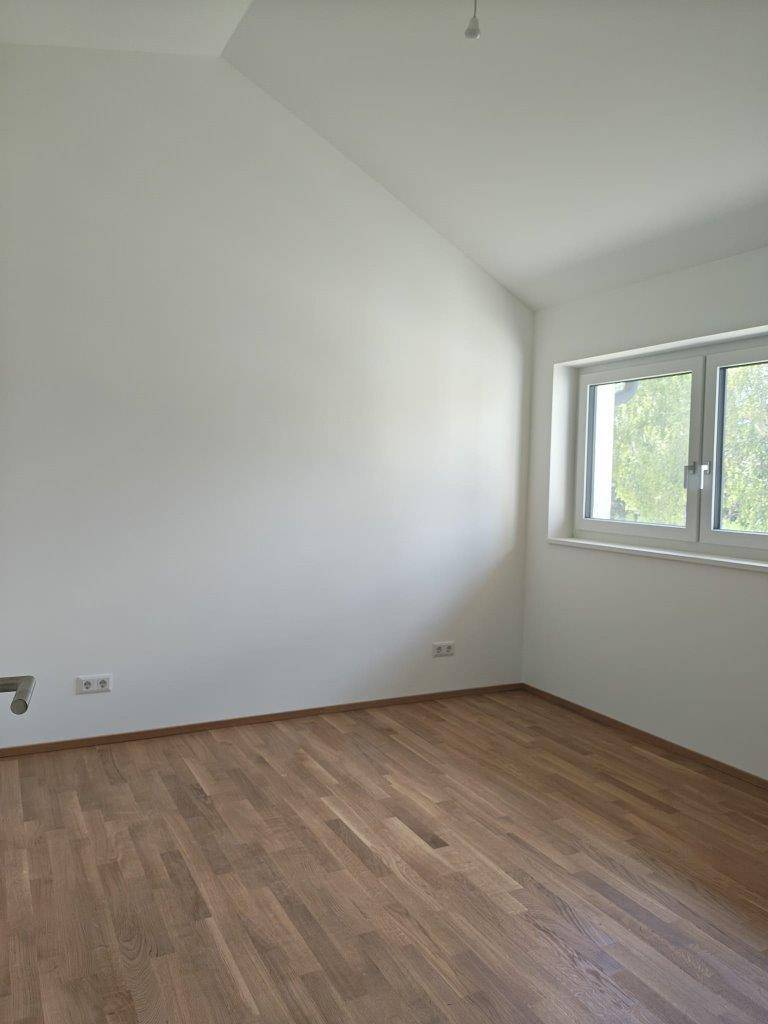 Unmöbliertes Schlafzimmer mit Holzboden, weiß gestrichenen Wänden und Fenster mit Blick ins Grüne.