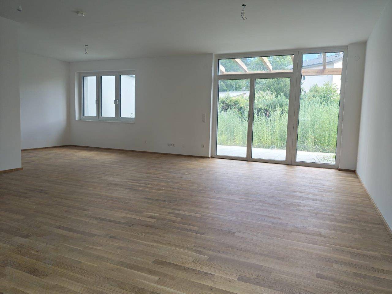 Leerer Wohnbereich mit Holzboden und großen Fenstern zum Garten, bereit zur individuellen Gestaltung.
