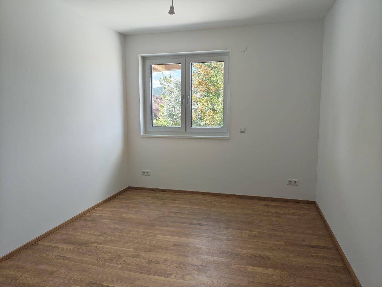Leeres Schlafzimmer mit Holzboden und Fenster, das viel Tageslicht hereinlässt und Ausblick bietet.