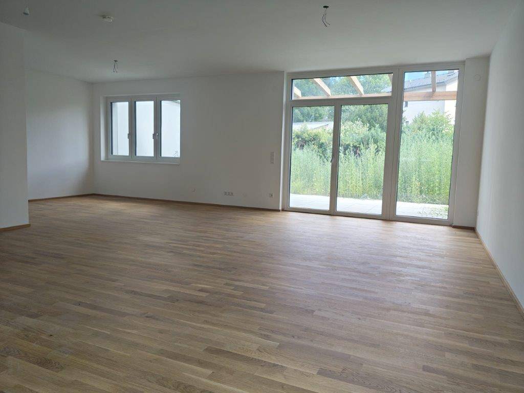 Heller, unmöblierter Wohnraum mit Holzboden und großen Fenstern zum Außenbereich.