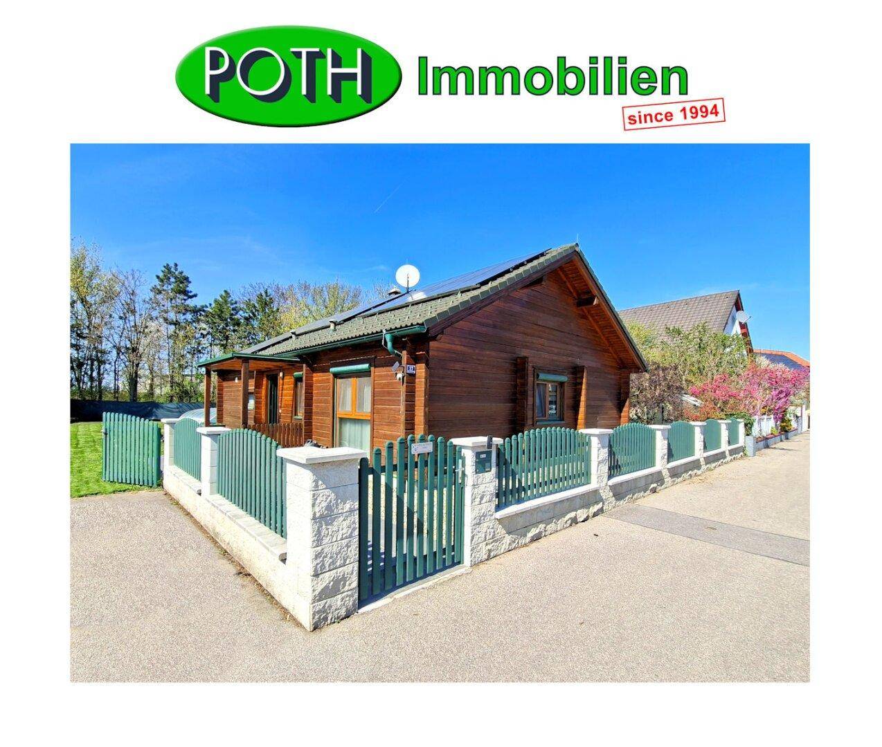 Ansicht des Holzhauses von der Straße mit grünem Zaun und Vorgarten.