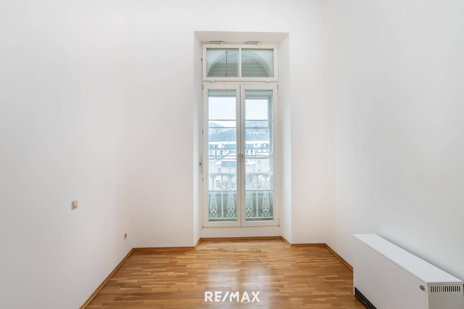 Helles Zimmer mit hohen Fenstern, Parkettboden und Blick nach draußen.