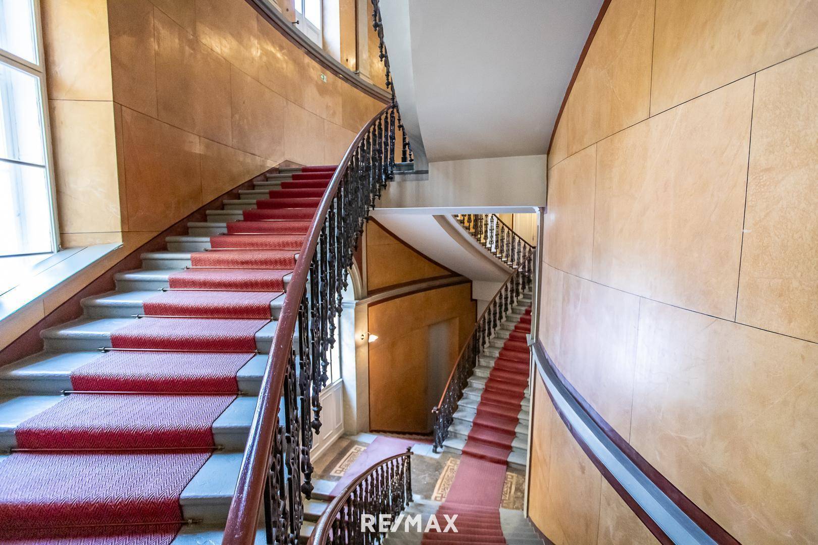 Elegantes Treppenhaus mit rotem Teppich, kunstvollem Geländer und hohen Wänden.