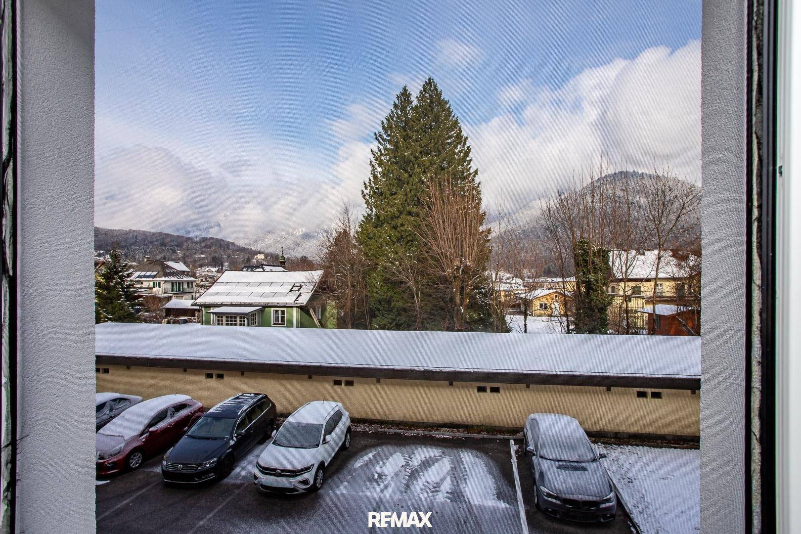 Blick aus dem Fenster auf einen verschneiten Parkplatz mit Bäumen und Bergen im Hintergrund.