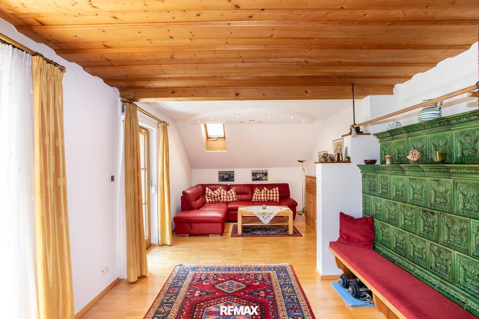 Offener Wohnbereich mit Holzbalkendecke, rotem Sofa und traditionellem Kachelofen.