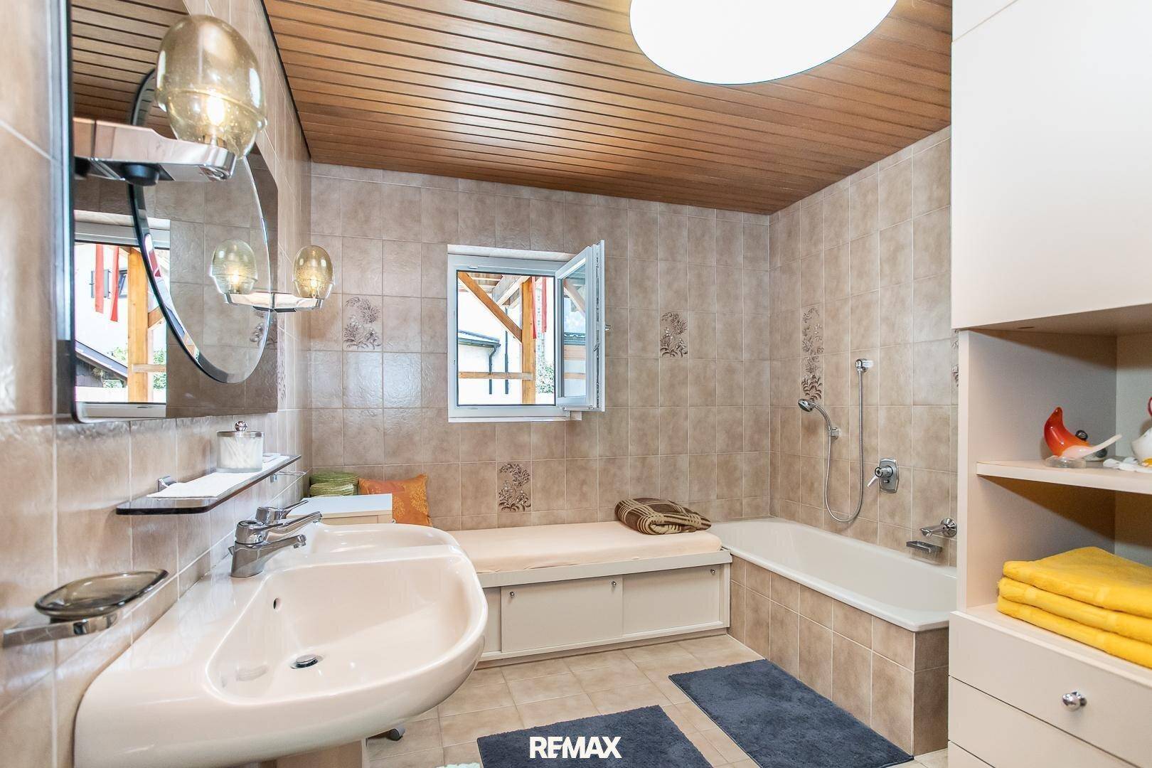 Gefliestes Badezimmer mit Badewanne, Waschbecken, Holzdecke und einem kleinen Fenster.