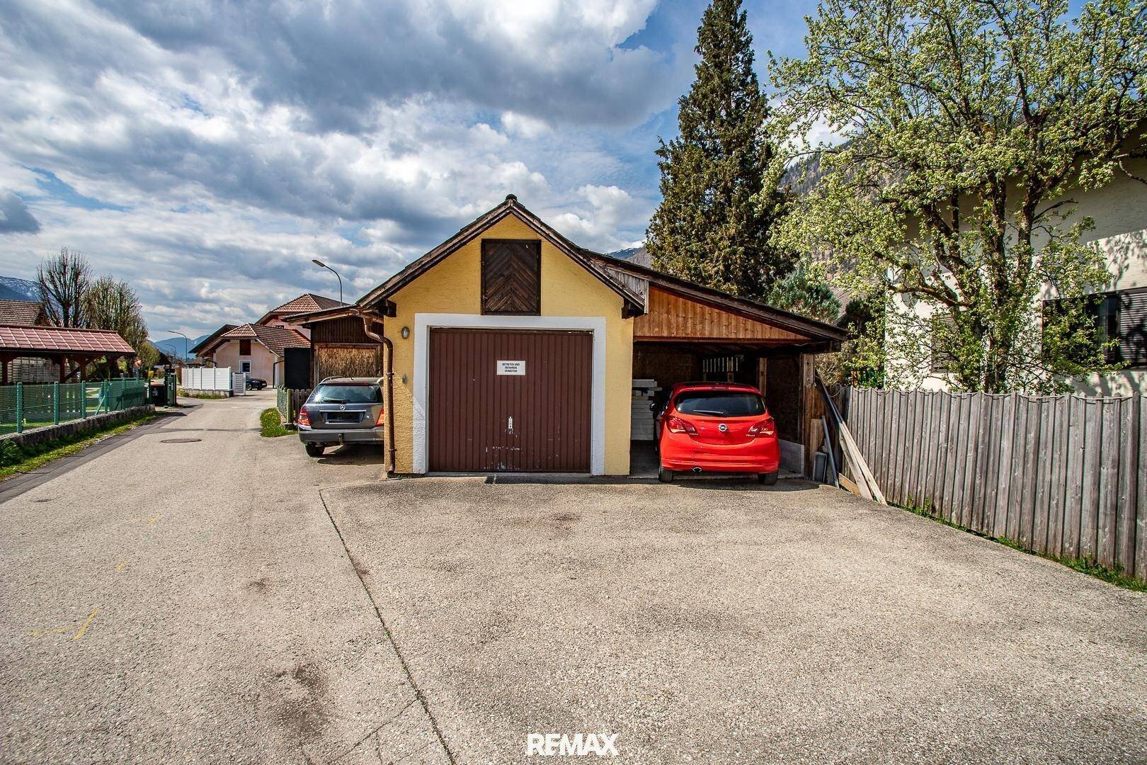 Gelbe Garage mit braunem Tor und Carport, umgeben von Bäumen und einer ländlichen Umgebung.