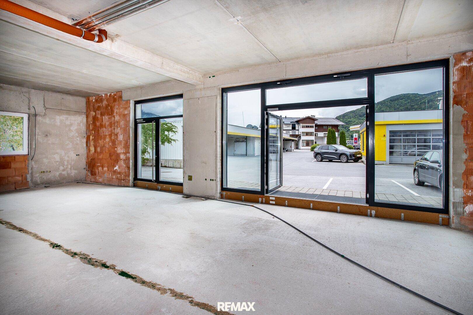 Innenansicht eines Rohbaus mit Betonboden und großen Glasfronten, die auf einen Parkplatz blicken.
