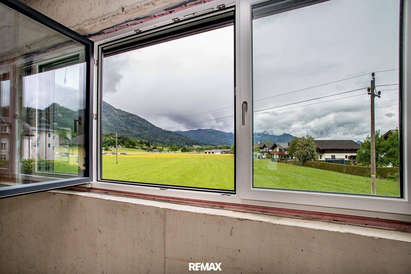 Fensterblick aus dem Innenraum auf grüne Wiesen und die umliegende Berglandschaft unter bewölktem Himmel.