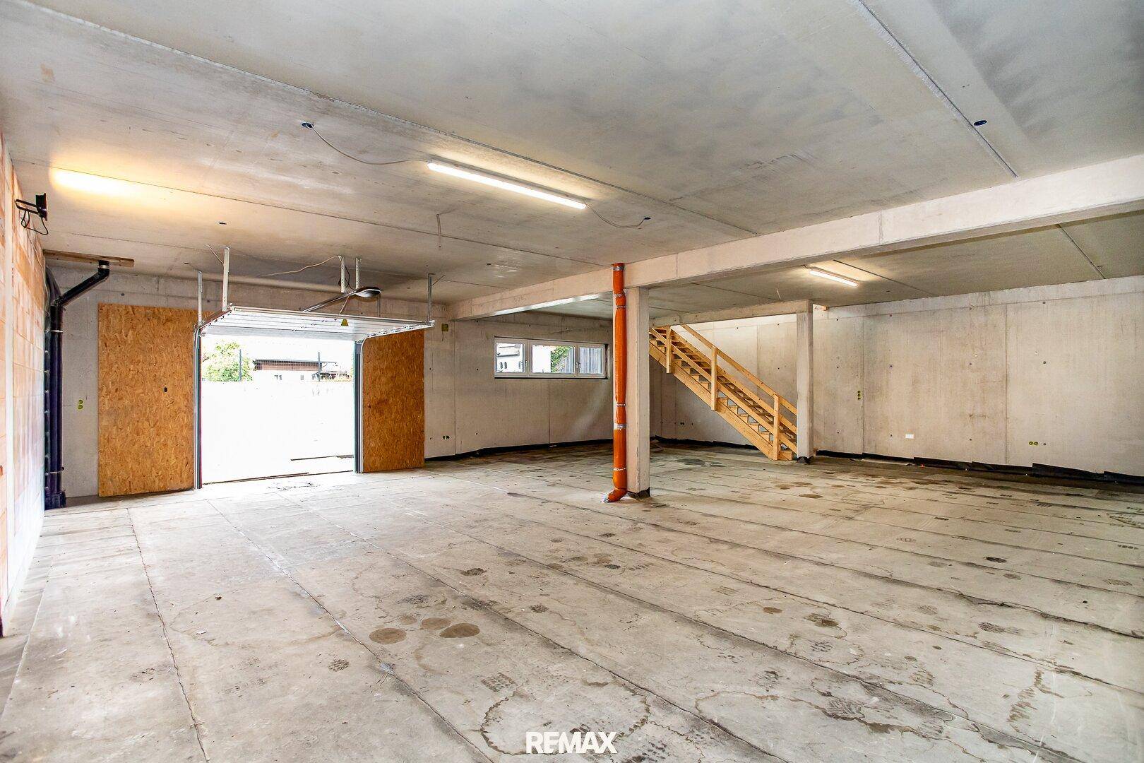 Weitläufiger Kellerraum mit offenem Garagentor, Betonboden und einer Holztreppe, die in die obere Etage führt.