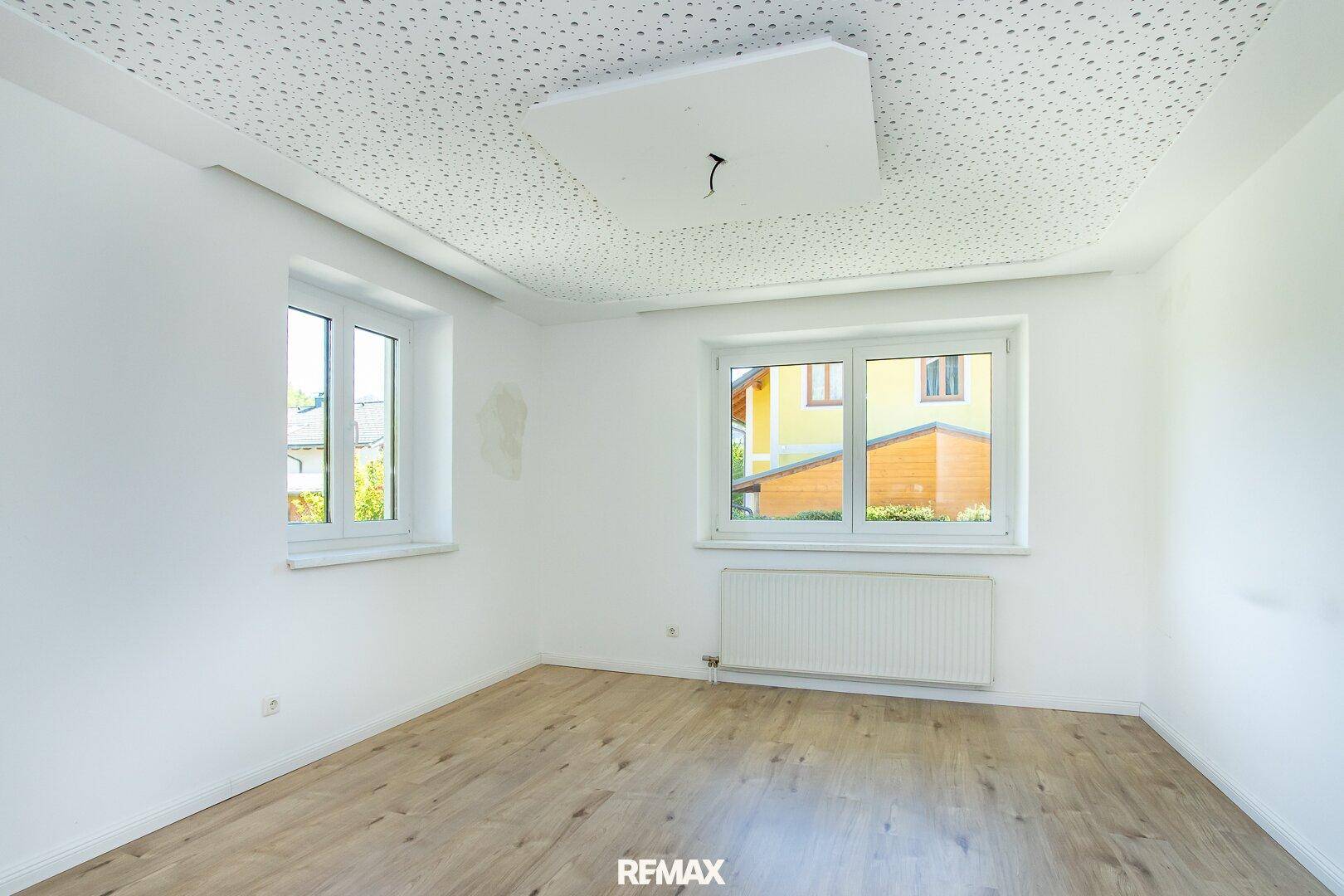 Freundliches Zimmer mit Laminatboden, zwei Fenstern und weiß gestrichenen Wänden.