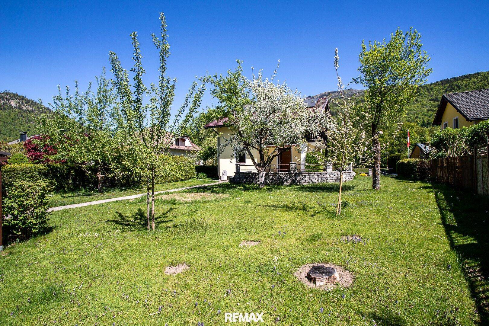 Weitläufiger Garten mit Rasen, jungen Bäumen und Blick auf das Haus und die Berge.