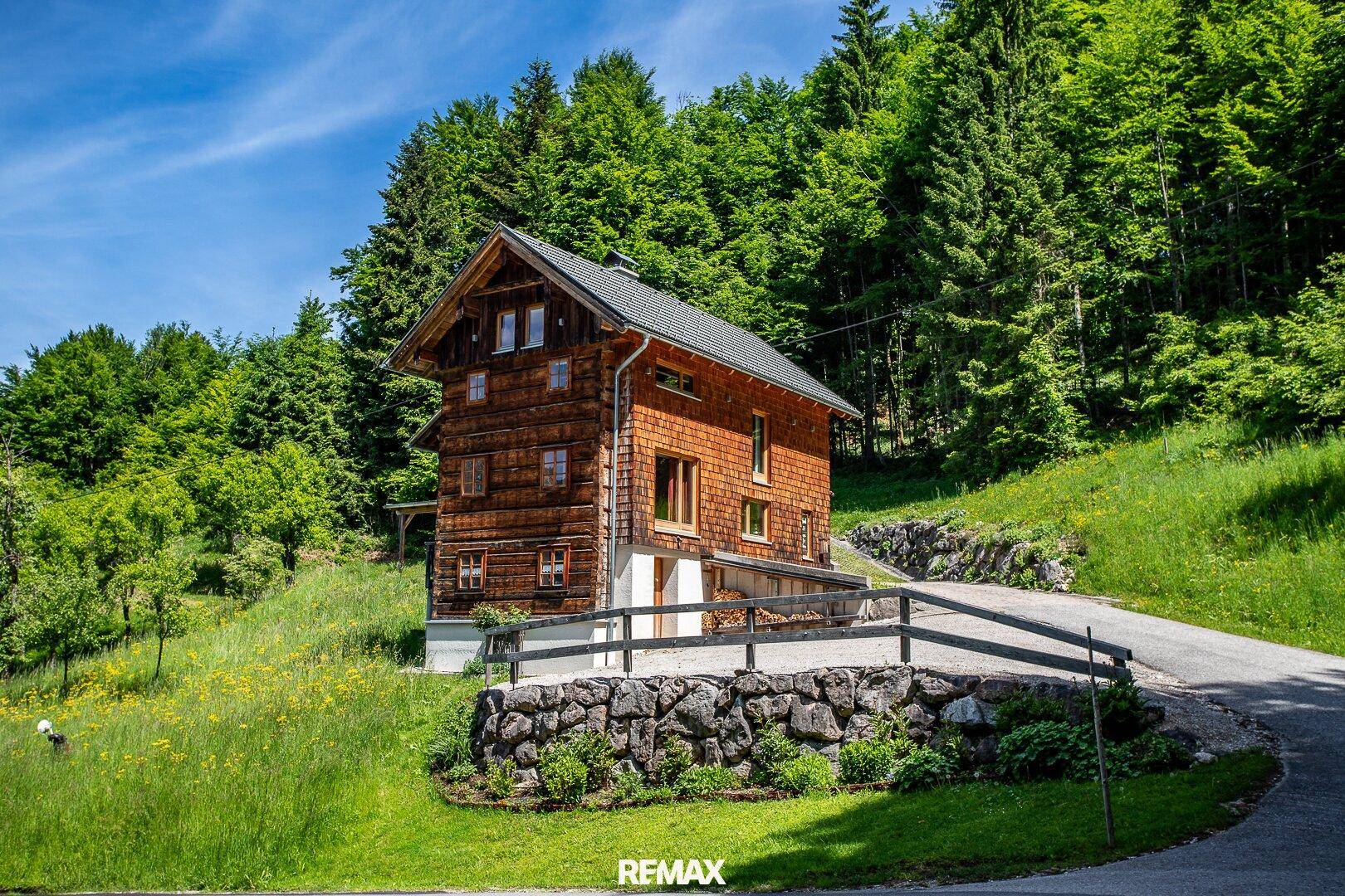Ein rustikales Holzhaus mit Satteldach, umgeben von grünen Hügeln und einem Wald im Hintergrund.