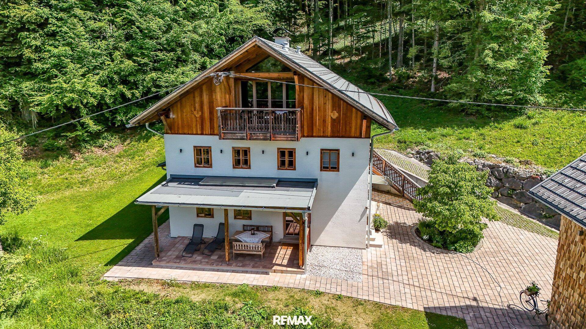 Traditionelles Haus mit weißer Fassade und Holzbalkon, umgeben von grüner Natur und Zufahrt.