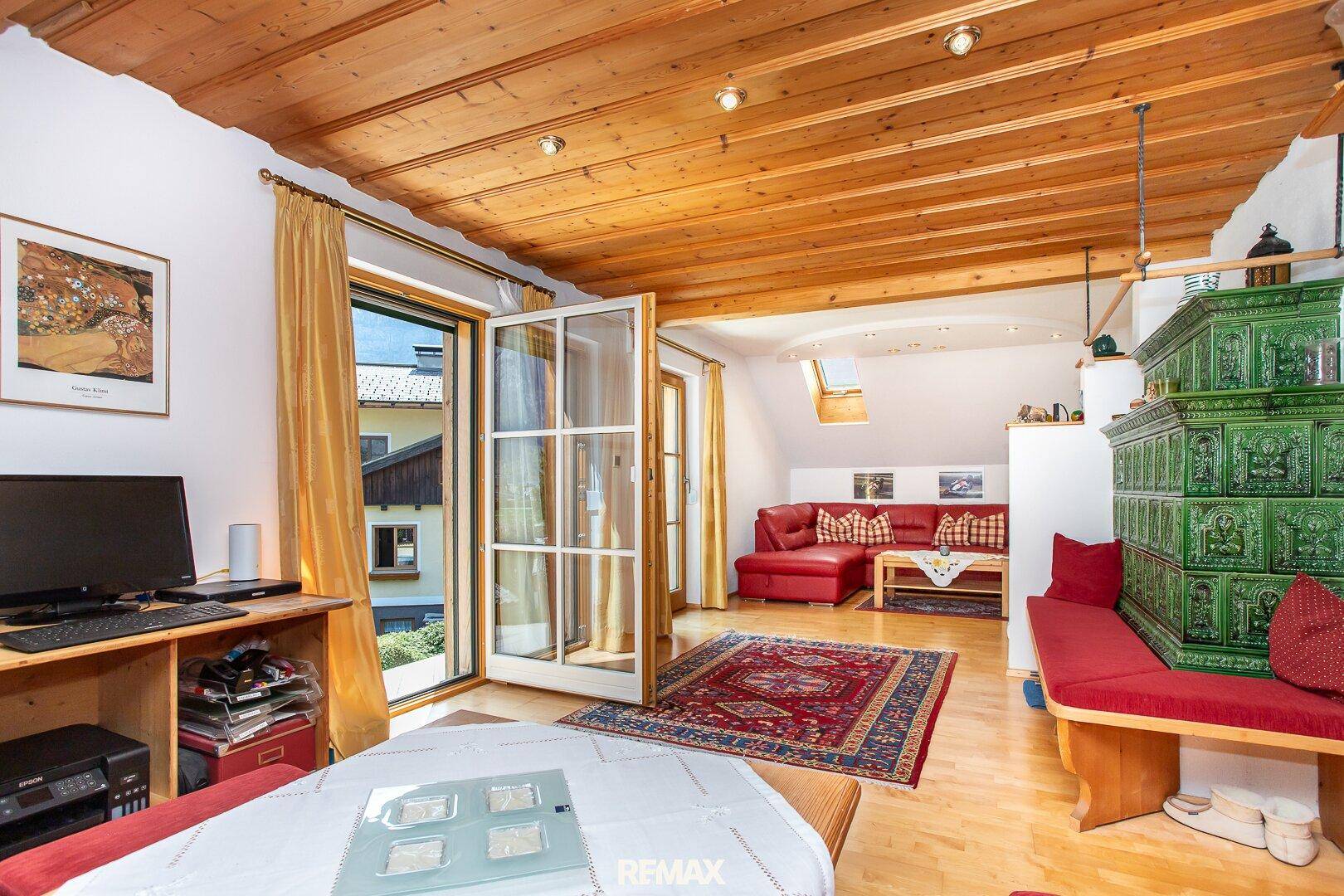 Gemütliches Wohnzimmer im alpinen Stil mit Holzbalkendecke, Kachelofen und Zugang zum Balkon.