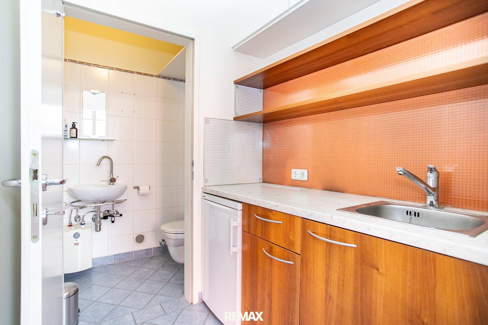 Kleine Teeküche mit Spüle, Kühlschrank und Holzschränken neben einem Badezimmer.
