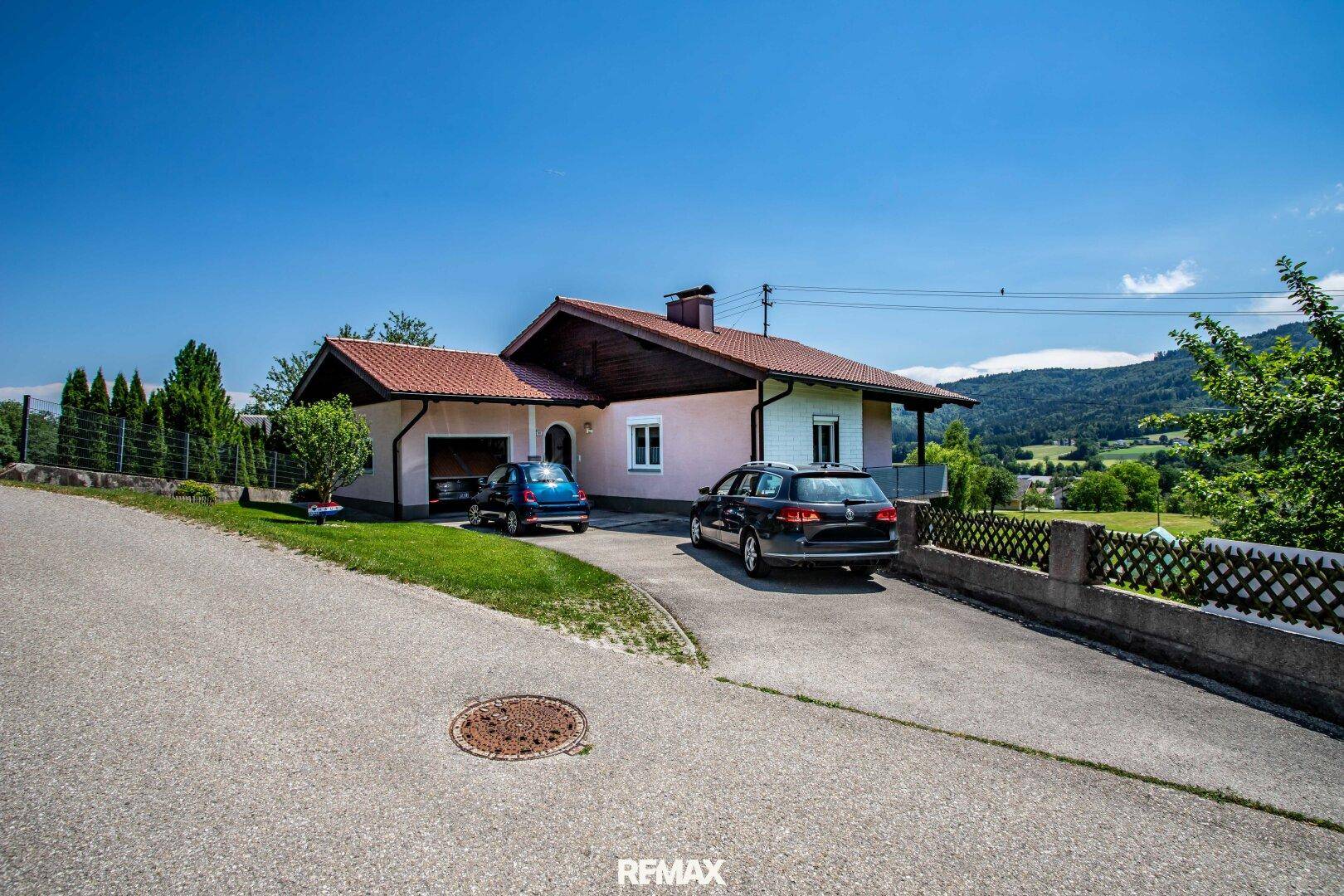 Vorderansicht des Hauses mit Einfahrt und geparkten Autos vor der grünen Hügellandschaft.