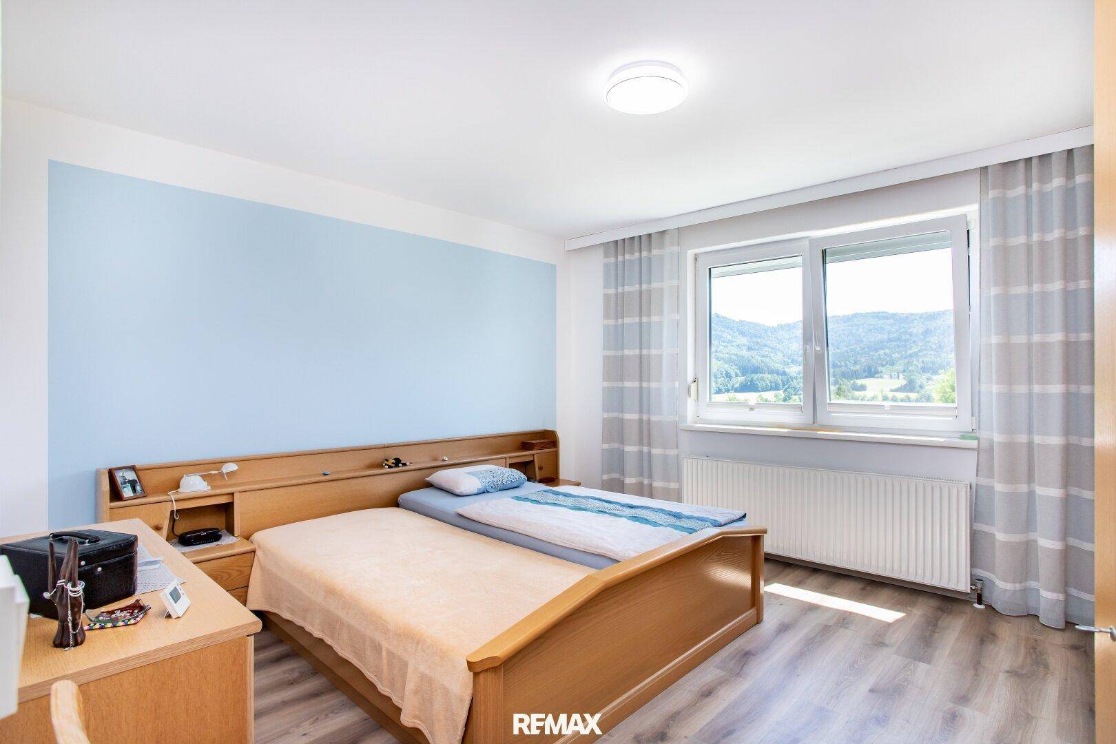 Schlafzimmer mit Doppelbett, blauer Akzentwand und Fenster mit Blick auf die Landschaft.