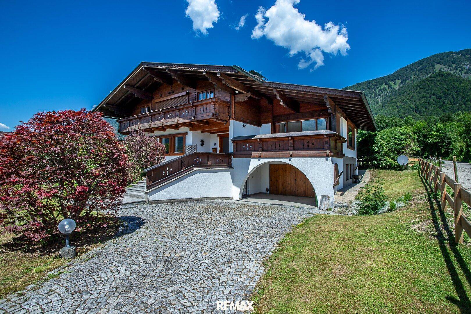 Charmantes Holzhaus mit gepflasterter Auffahrt und traditioneller Architektur vor alpiner Kulisse.