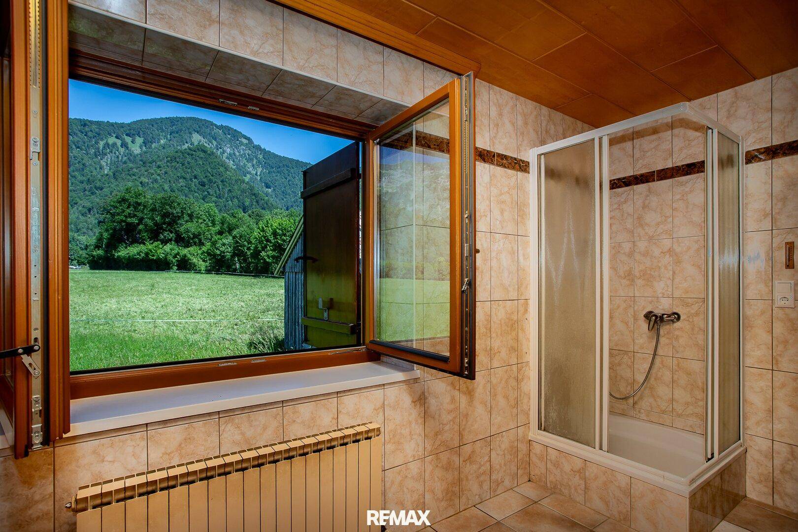 Helles Badezimmer mit Fliesen, Duschkabine und Fenster mit weitem Blick ins Grüne und auf die Berge.