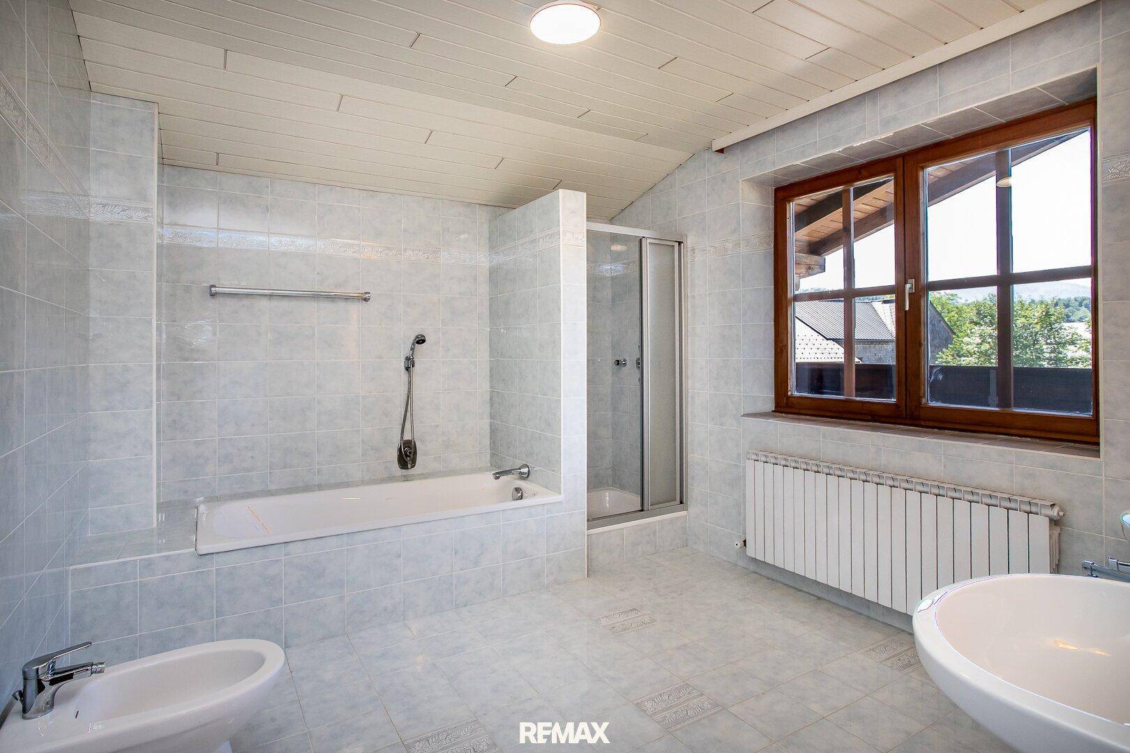 Geräumiges Badezimmer mit hellgrauen Fliesen, Badewanne, Dusche, Bidet und Fenster.