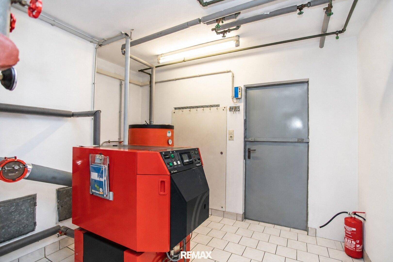 Heizungsraum mit großem roten Heizkessel und sichtbaren Rohrleitungen auf Fliesenboden.