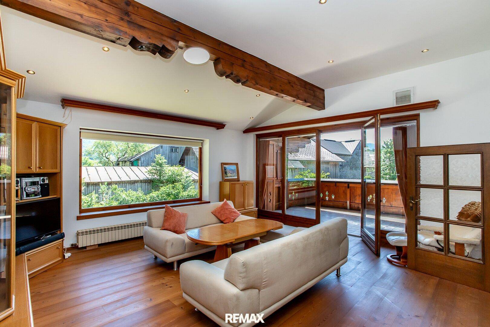 Gemütliches Wohnzimmer mit Holzboden, sichtbaren Holzbalken und Zugang zu einem sonnigen Balkon.