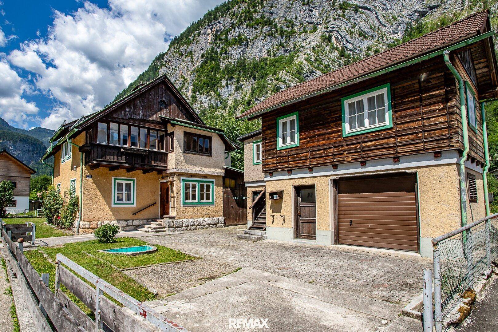 Zwei traditionelle Häuser mit Garagenzufahrt und alpiner Architektur in bergiger Landschaft.