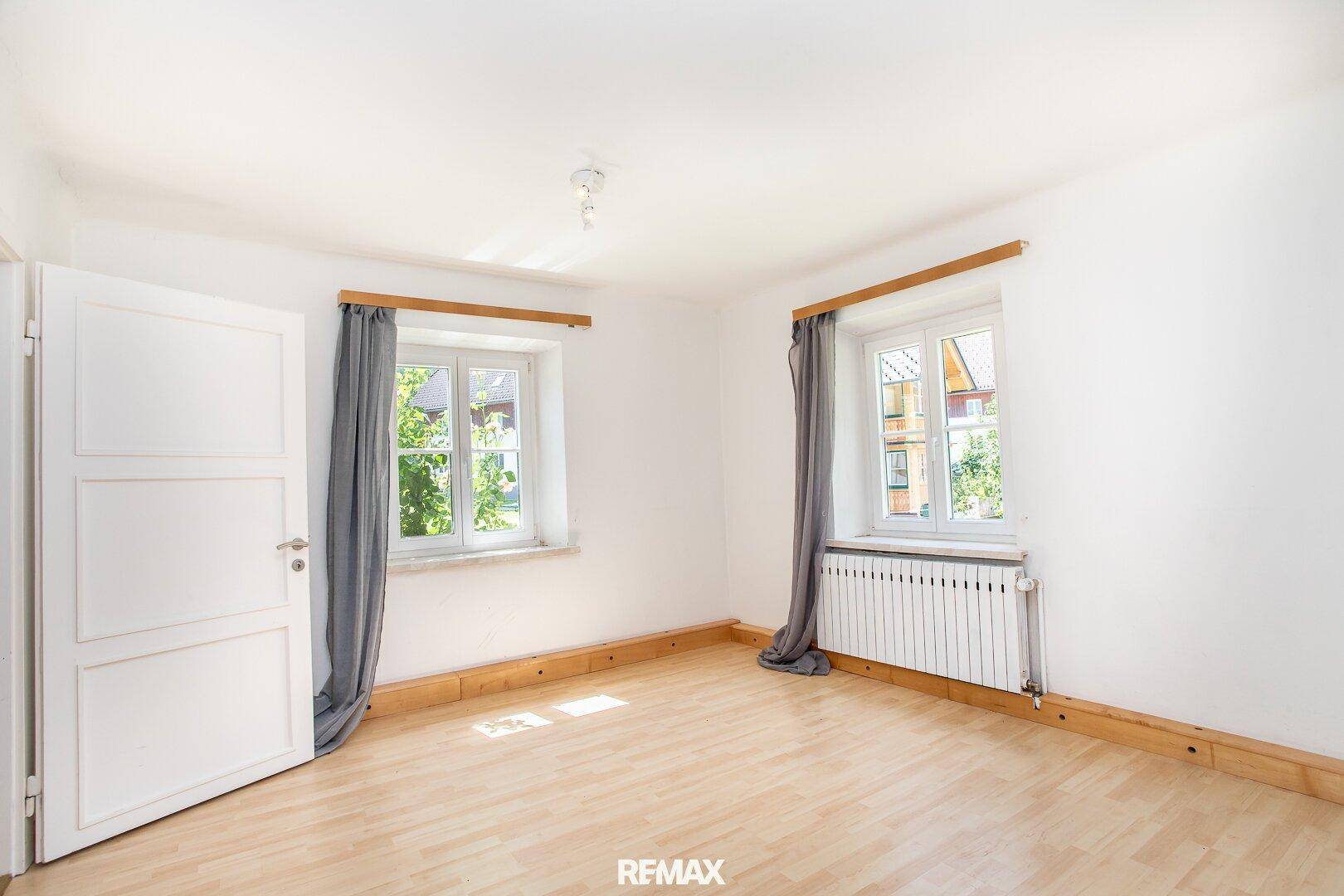 Helles Schlafzimmer mit Holzboden, weißen Wänden und zwei Fenstern mit Blick ins Grüne.