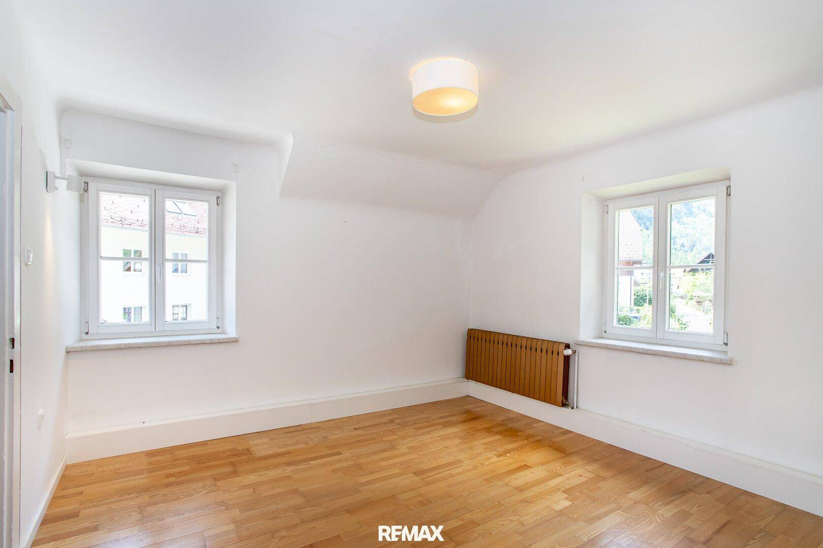 Helles Schlafzimmer mit Holzboden, weißen Wänden und zwei Fenstern, die viel Licht hereinlassen.