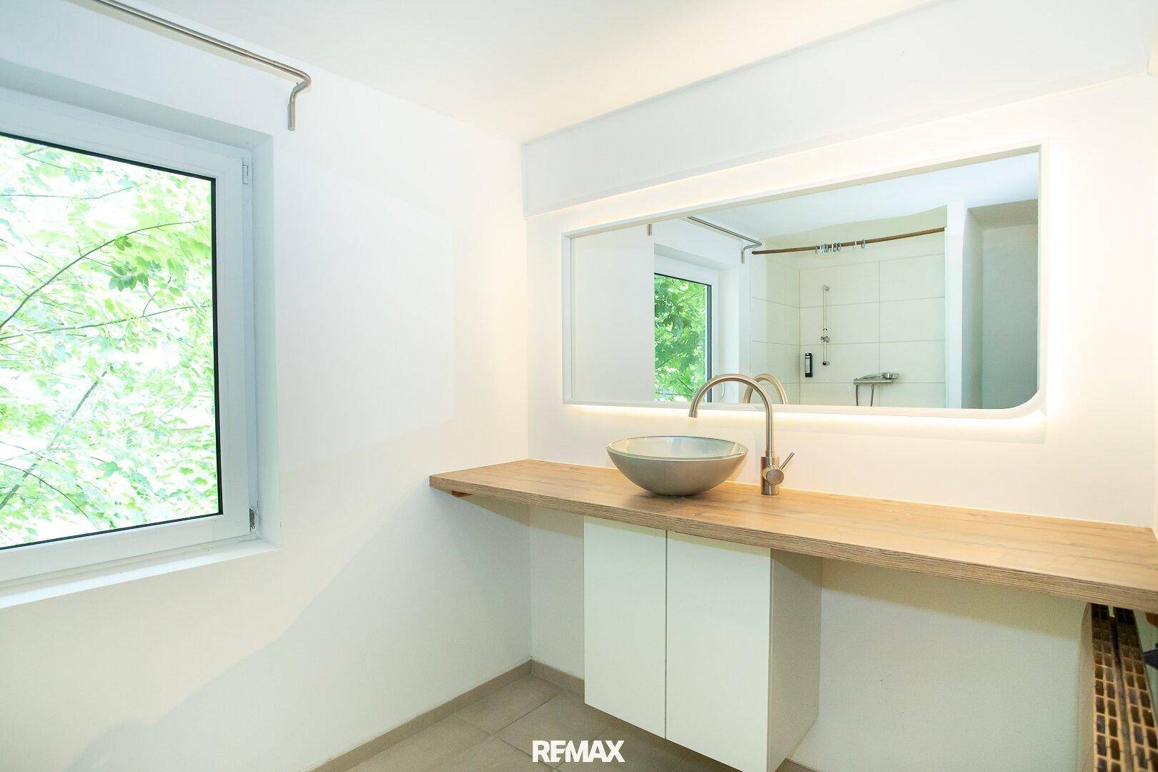 Modernes Badezimmer mit stilvollem Waschbecken, großem Spiegel und Fenster mit Blick ins Grüne.
